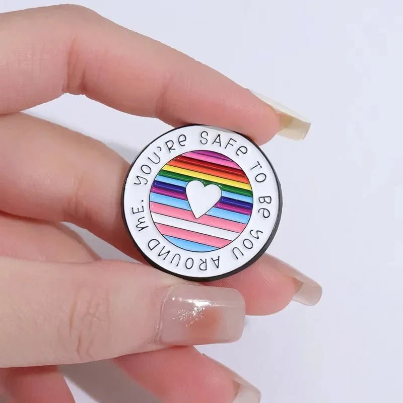 A hand holds an enamel pin featuring a heart atop rainbow pride flag stripes. The text around the edge reads, You’re safe to be you around me.