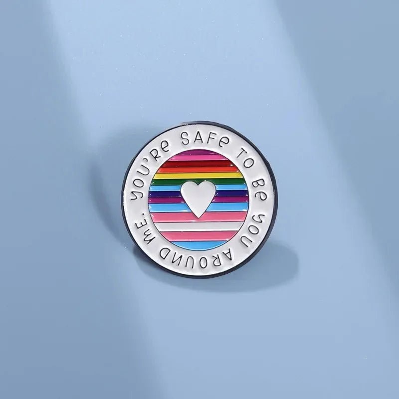 A round enamel pin features rainbow pride flag stripes, a white heart at the center, and YOURE SAFE TO BE YOU AROUND ME around the edge, displayed on a light blue background.