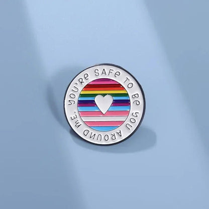 A round enamel pin features rainbow pride flag stripes, a white heart at the center, and YOURE SAFE TO BE YOU AROUND ME around the edge, displayed on a light blue background.