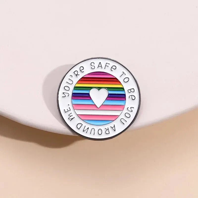 round enamel pin features a heart at the center over rainbow pride stripes, with Youre safe to be you around me around the edge. It’s displayed on a soft, light-colored background.
