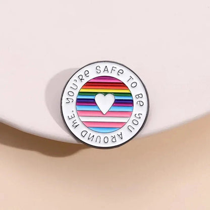 round enamel pin features a heart at the center over rainbow pride stripes, with Youre safe to be you around me around the edge. It’s displayed on a soft, light-colored background.