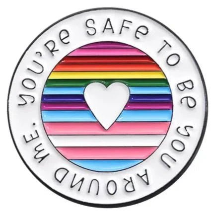 enamel pin features a circular design with a cut-out heart center, rainbow pride stripes, and the phrase YOURE SAFE TO BE YOU AROUND ME around the edge.