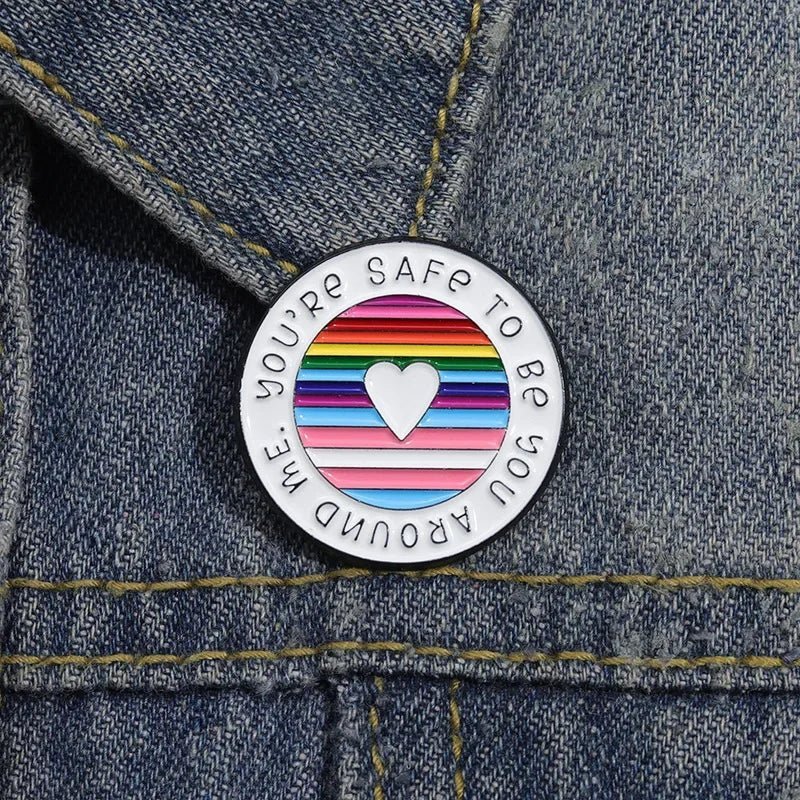 round enamel pin on denim shows a white heart over rainbow pride stripes, with You’re safe to be you around me around the edge.